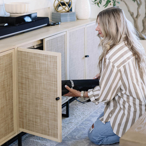 Woman opening a cabinet in a modern living room to stow away rolled up puzzle mat with a painting and plant in the background.